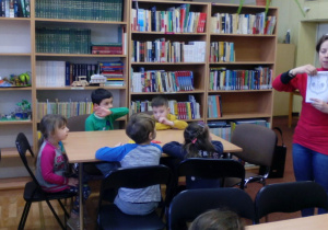 Dzieci siedzące przy stolikach. Prowadząca zajęcia pokazuje kolorowankę herbu miasta Łask.